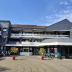 Modernes Gebäude mit Balkonen, blauem Himmel und Sitzgelegenheiten im Freien an einem sonnigen Tag.