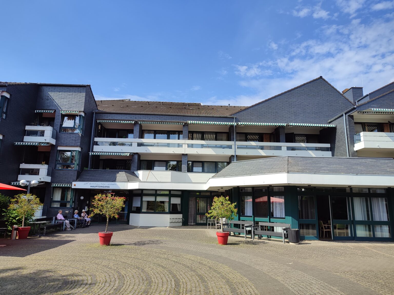 Modernes Gebäude mit Balkonen, blauem Himmel und Sitzgelegenheiten im Freien an einem sonnigen Tag.