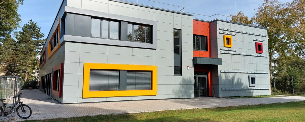 Modernes Gebäude mit bunten Akzenten, umgeben von Bäumen und Grünfläche unter klarem Himmel.