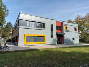 Modernes Gebäude mit bunten Akzenten, umgeben von Bäumen und Grünfläche unter klarem Himmel.