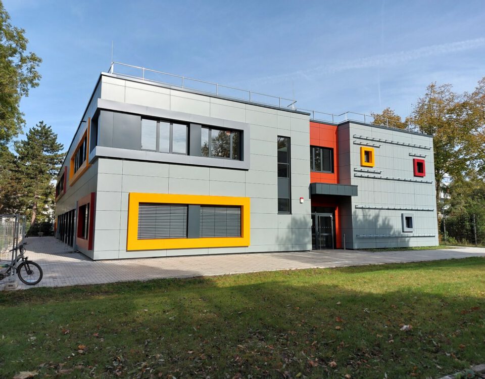 Modernes Gebäude mit bunten Akzenten, umgeben von Bäumen und Grünfläche unter klarem Himmel.