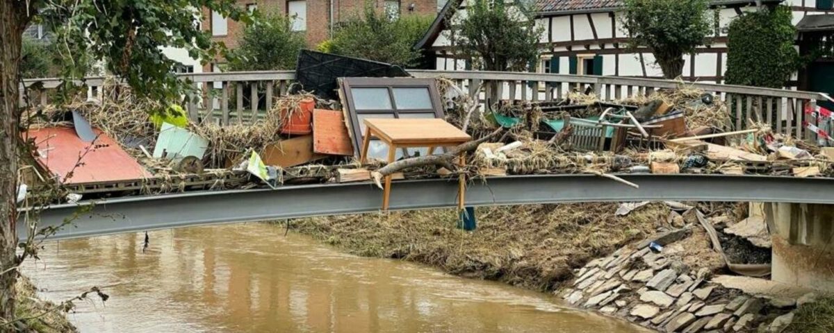 Brücke mit Überschwemmungsschäden und Trümmern, traditionelle Häuser im Hintergrund, Naturkatastrophe in einem Dorf.