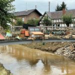 Brücke mit Überschwemmungsschäden und Trümmern, traditionelle Häuser im Hintergrund, Naturkatastrophe in einem Dorf.