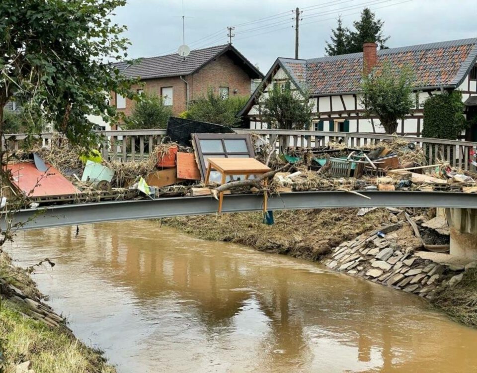 Brücke mit Überschwemmungsschäden und Trümmern, traditionelle Häuser im Hintergrund, Naturkatastrophe in einem Dorf.