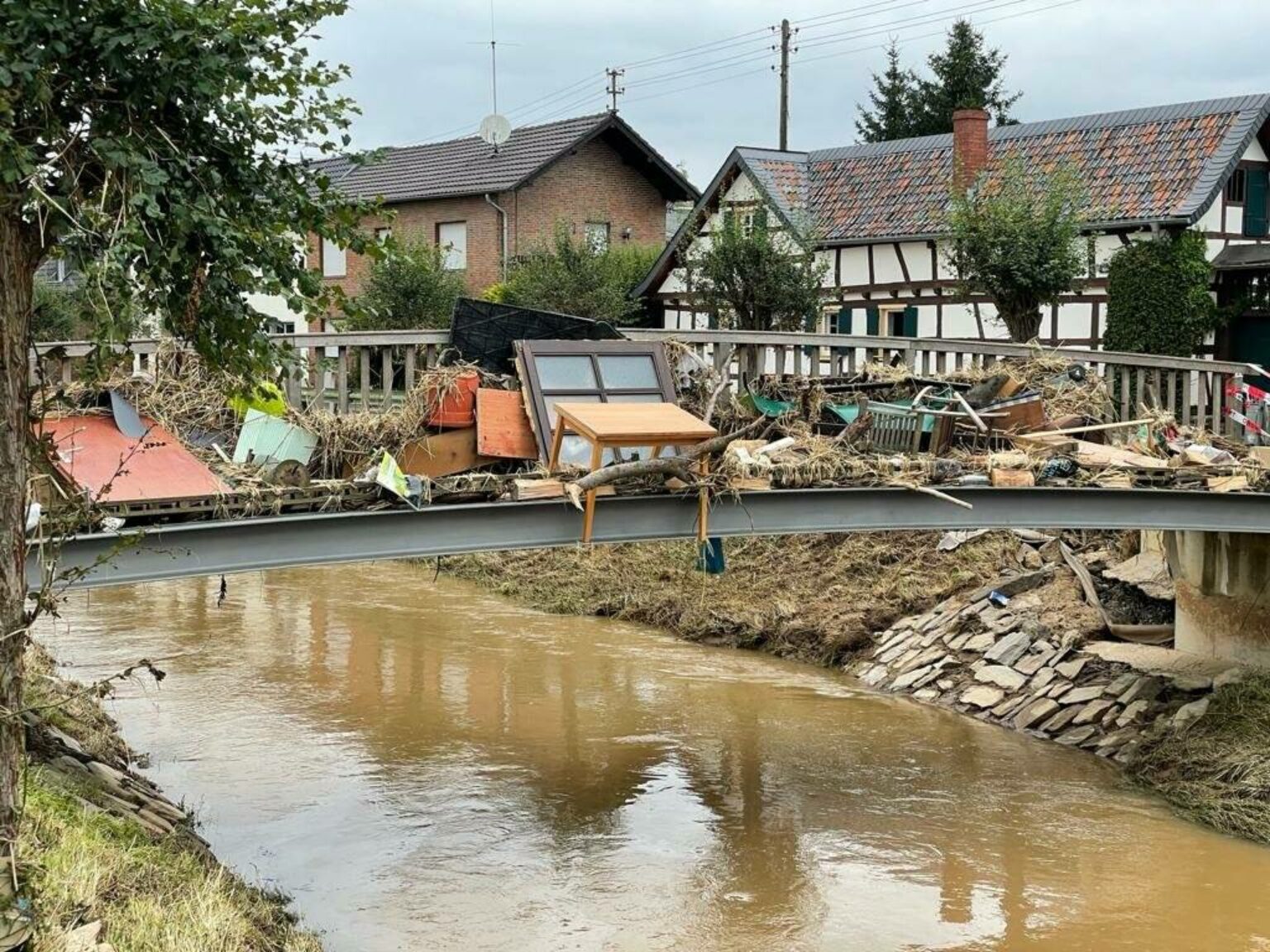 Brücke mit Überschwemmungsschäden und Trümmern, traditionelle Häuser im Hintergrund, Naturkatastrophe in einem Dorf.
