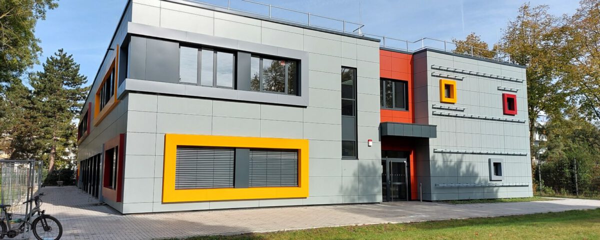 Modernes Gebäude mit bunter Fassade und großen Fenstern, umgeben von Bäumen und Rasen, an sonnigem Tag.