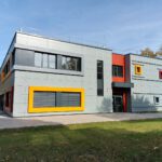 Modernes Gebäude mit bunter Fassade und großen Fenstern, umgeben von Bäumen und Rasen, an sonnigem Tag.