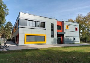 Modernes Gebäude mit bunter Fassade und großen Fenstern, umgeben von Bäumen und Rasen, an sonnigem Tag.