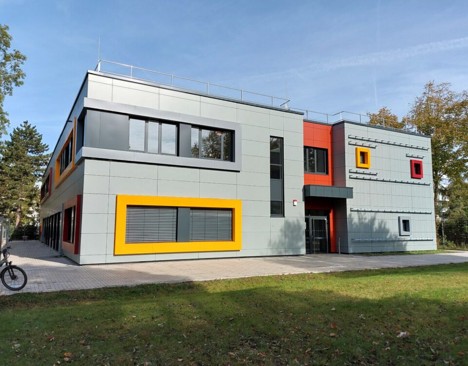 Modernes Gebäude mit bunter Fassade und großen Fenstern, umgeben von Bäumen und Rasen, an sonnigem Tag.