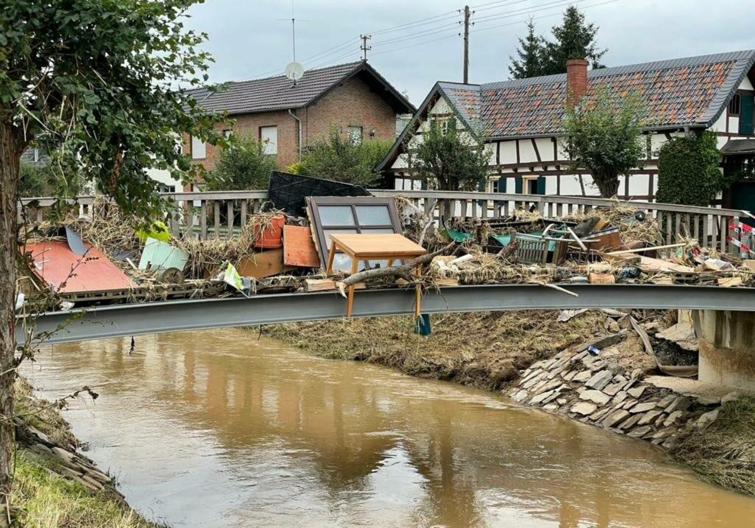 Zerstörte Brücke mit Treibgut nach Hochwasser in einem Dorf, zerstörte Häuser im Hintergrund.
