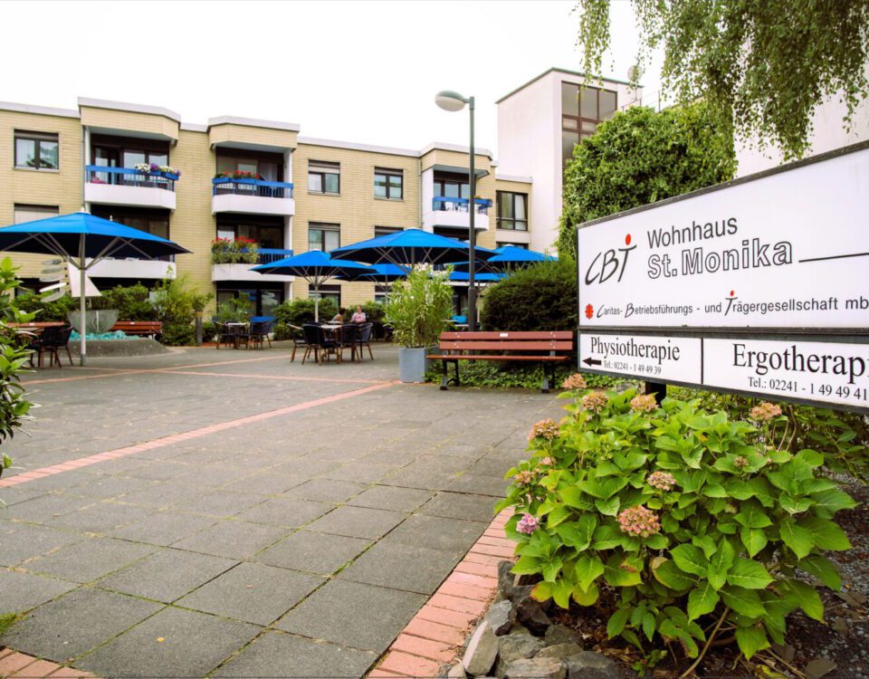 CBT Wohnhaus St. Monika mit blauen Sonnenschirmen, Hinweisschild zu Physiotherapie und Ergotherapie in Köln.