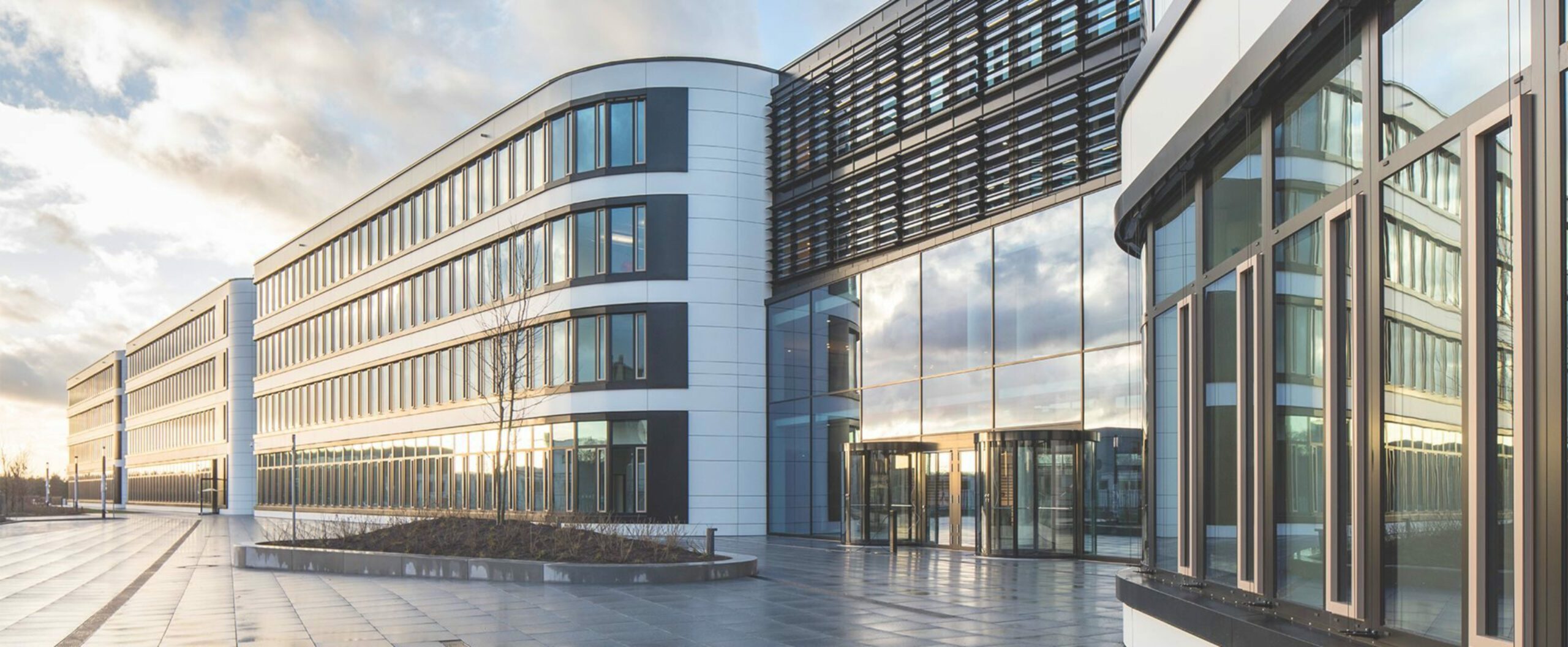 Modernes Bürogebäude mit Glasfassade und reflektierendem Himmel in urbaner Umgebung.