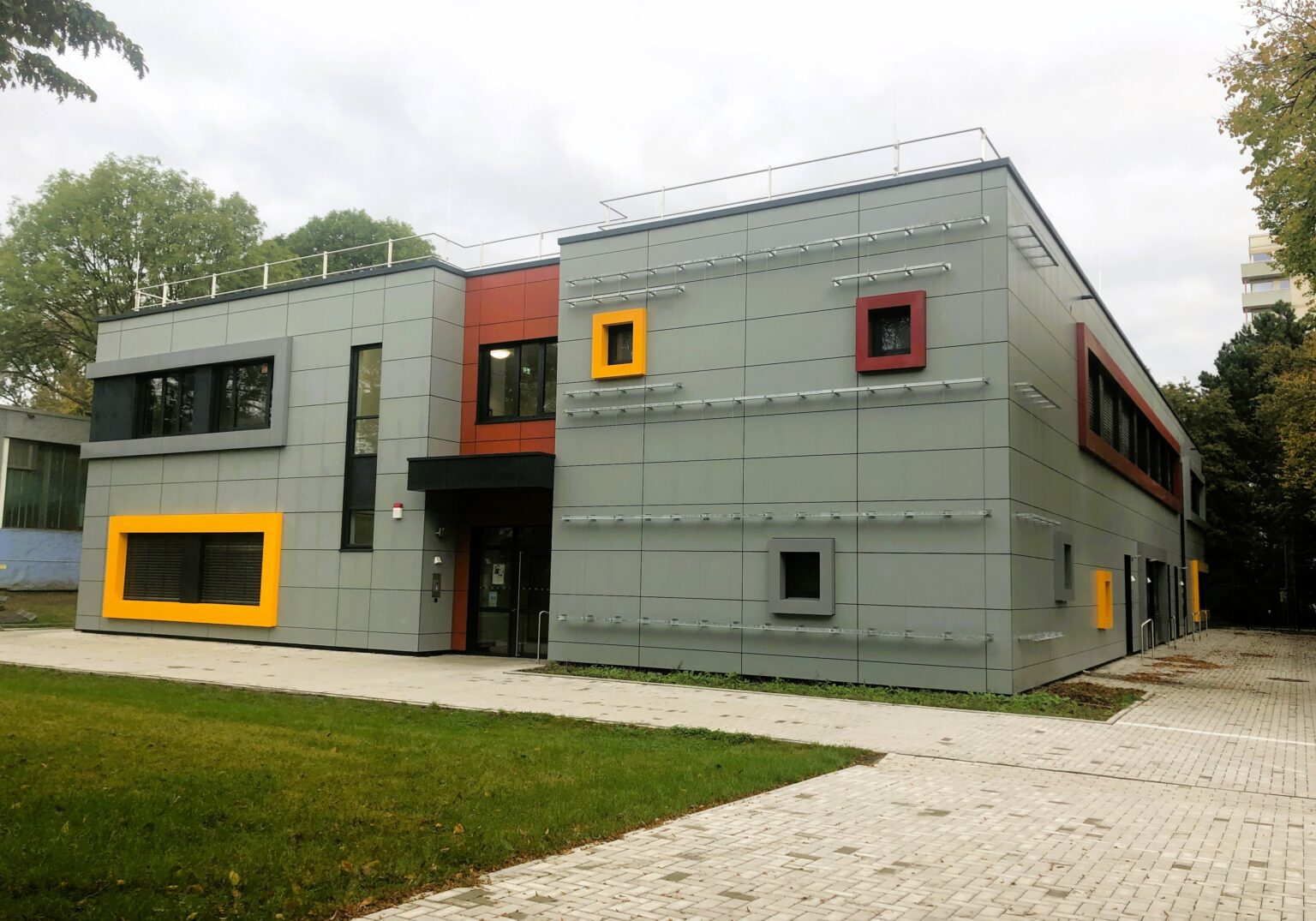 Modernes Gebäude mit bunten Fensterrändern und grauer Fassade, umgeben von Grünfläche, hellem Himmel im Hintergrund.