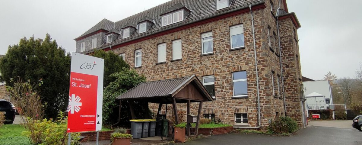 Altes Steingebäude des Wohnhauses St. Josef mit Schild und gepflegter Umgebung, bewölkter Himmel im Hintergrund.