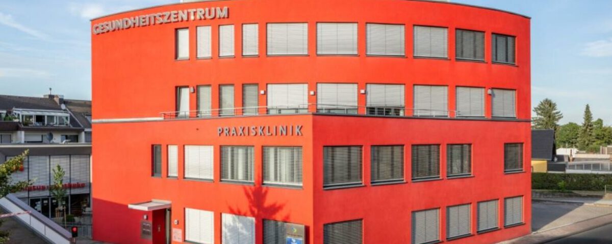 Rotes Gesundheitszentrum in moderner Architektur, Außenansicht bei Tageslicht, mit Beschriftung „Praxisklinik“.