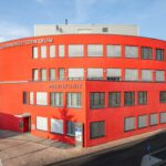 Rotes Gesundheitszentrum in moderner Architektur, Außenansicht bei Tageslicht, mit Beschriftung „Praxisklinik“.