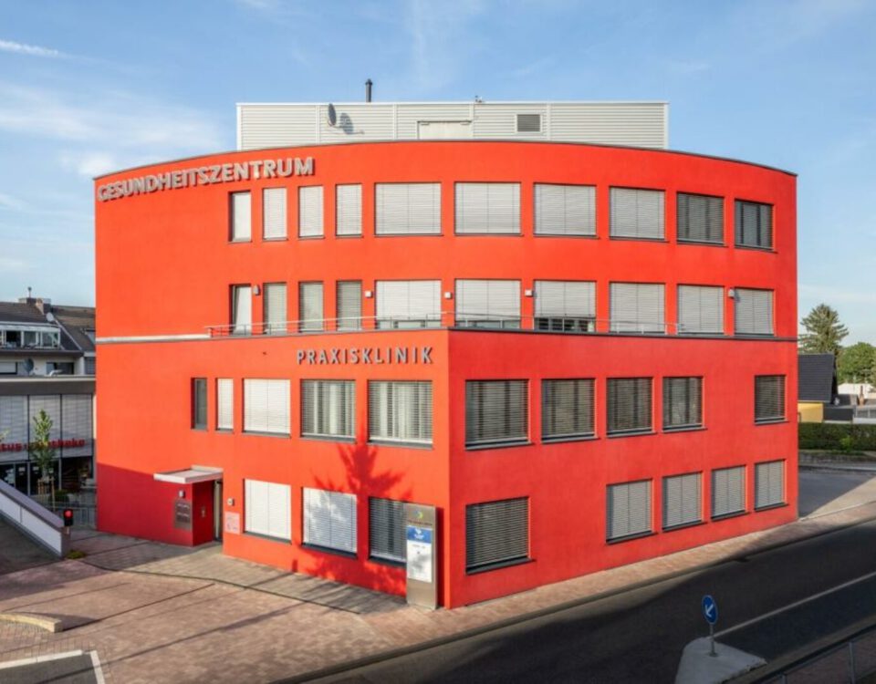 Rotes Gesundheitszentrum in moderner Architektur, Außenansicht bei Tageslicht, mit Beschriftung „Praxisklinik“.