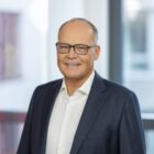 C9A3159_Favorit_AW_hochaufloesend-480x0-c-default Geschäftsmann in Anzug und Brille lächelt in modernem Büro, mit Fenstern und Kunstwerk im Hintergrund.