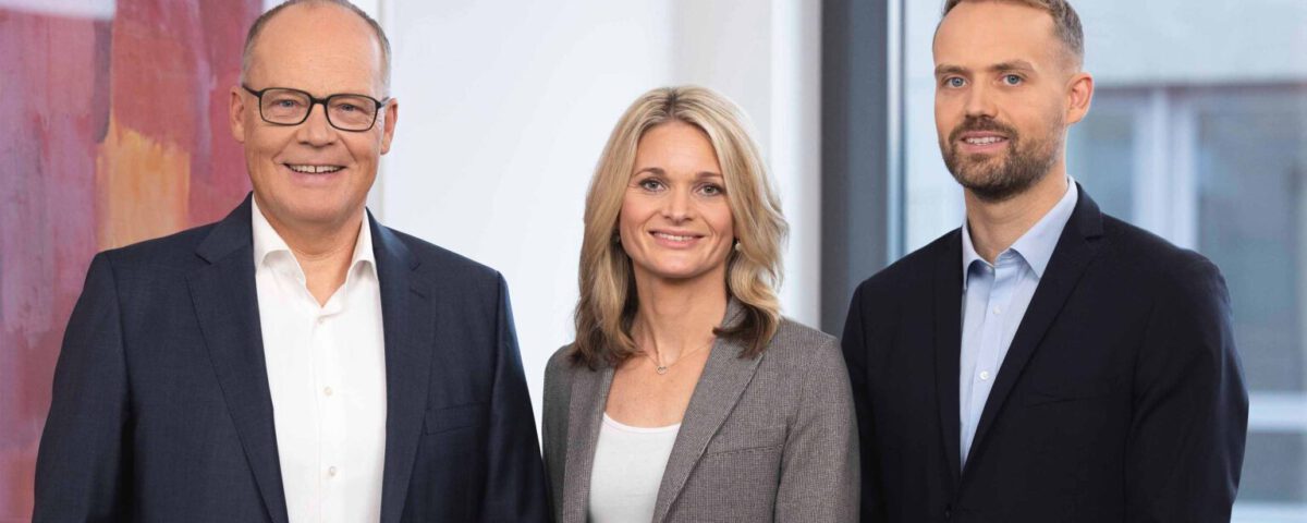 Drei Geschäftspersonen in formeller Kleidung lächeln vor einem Bürohintergrund in die Kamera.