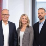 Drei Geschäftspersonen in formeller Kleidung lächeln vor einem Bürohintergrund in die Kamera.