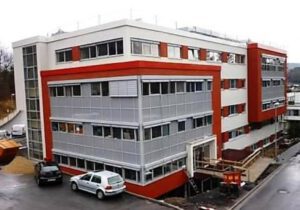 Modernes Bürogebäude mit rotem und grauem Fassadendesign, umgeben von Autos auf dem Parkplatz.