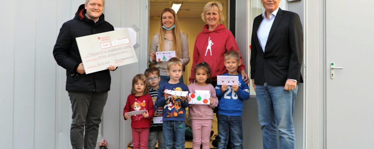 Gruppe überreicht Scheck an Kindereinrichtung vor Gebäude, Erwachsene und Kinder mit Zeichnungen stehen zusammen.