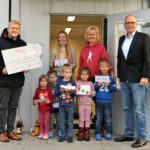 Gruppe überreicht Scheck an Kindereinrichtung vor Gebäude, Erwachsene und Kinder mit Zeichnungen stehen zusammen.