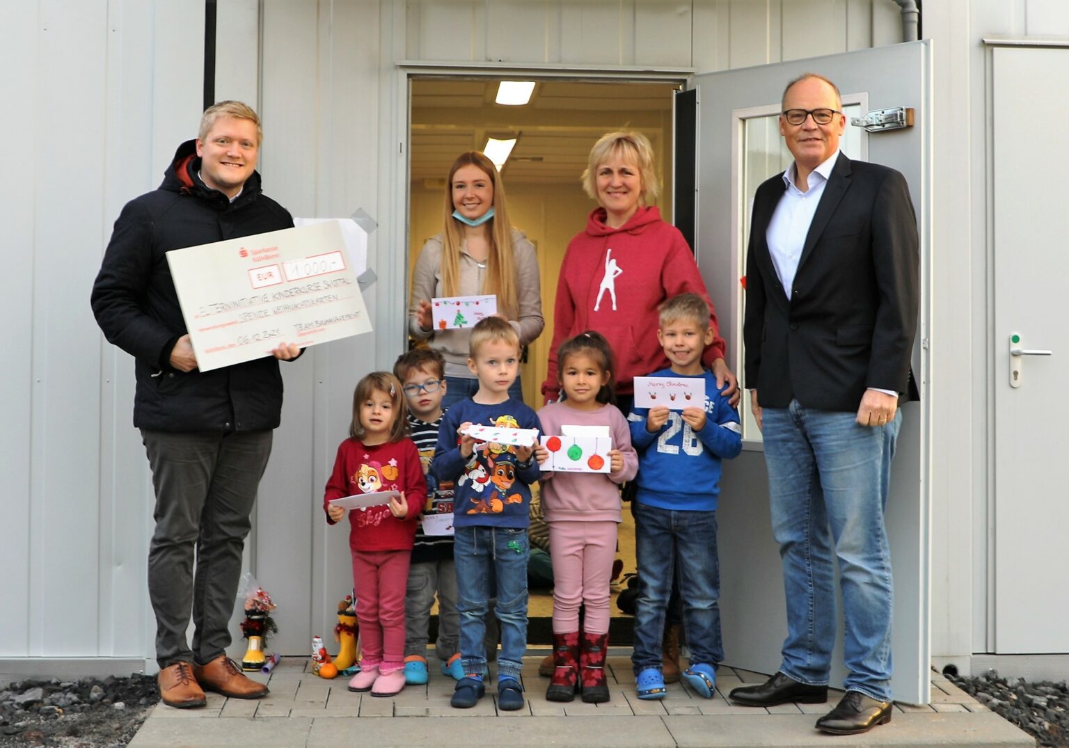 Personen überreichen Scheck an Kindergruppe vor einem Gebäude. Kinder halten Weihnachtskarten und lächeln in die Kamera.