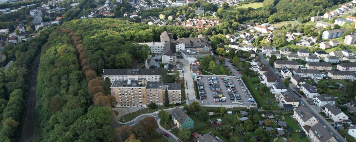 Luftaufnahme einer Stadtlandschaft mit Gebäuden, einem Parkplatz und einem bewaldeten Gebiet im Hintergrund.