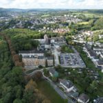 Luftaufnahme einer Stadtlandschaft mit Gebäuden, einem Parkplatz und einem bewaldeten Gebiet im Hintergrund.