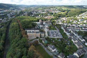 Luftaufnahme einer Stadtlandschaft mit Gebäuden, einem Parkplatz und einem bewaldeten Gebiet im Hintergrund.