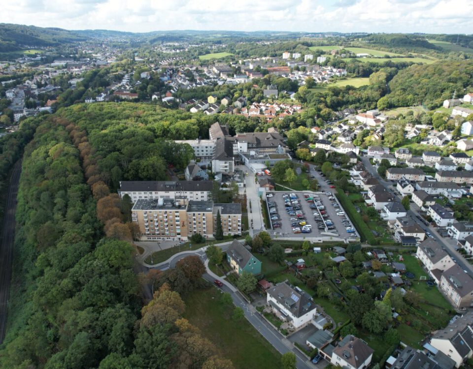 Luftaufnahme einer Stadtlandschaft mit Gebäuden, einem Parkplatz und einem bewaldeten Gebiet im Hintergrund.