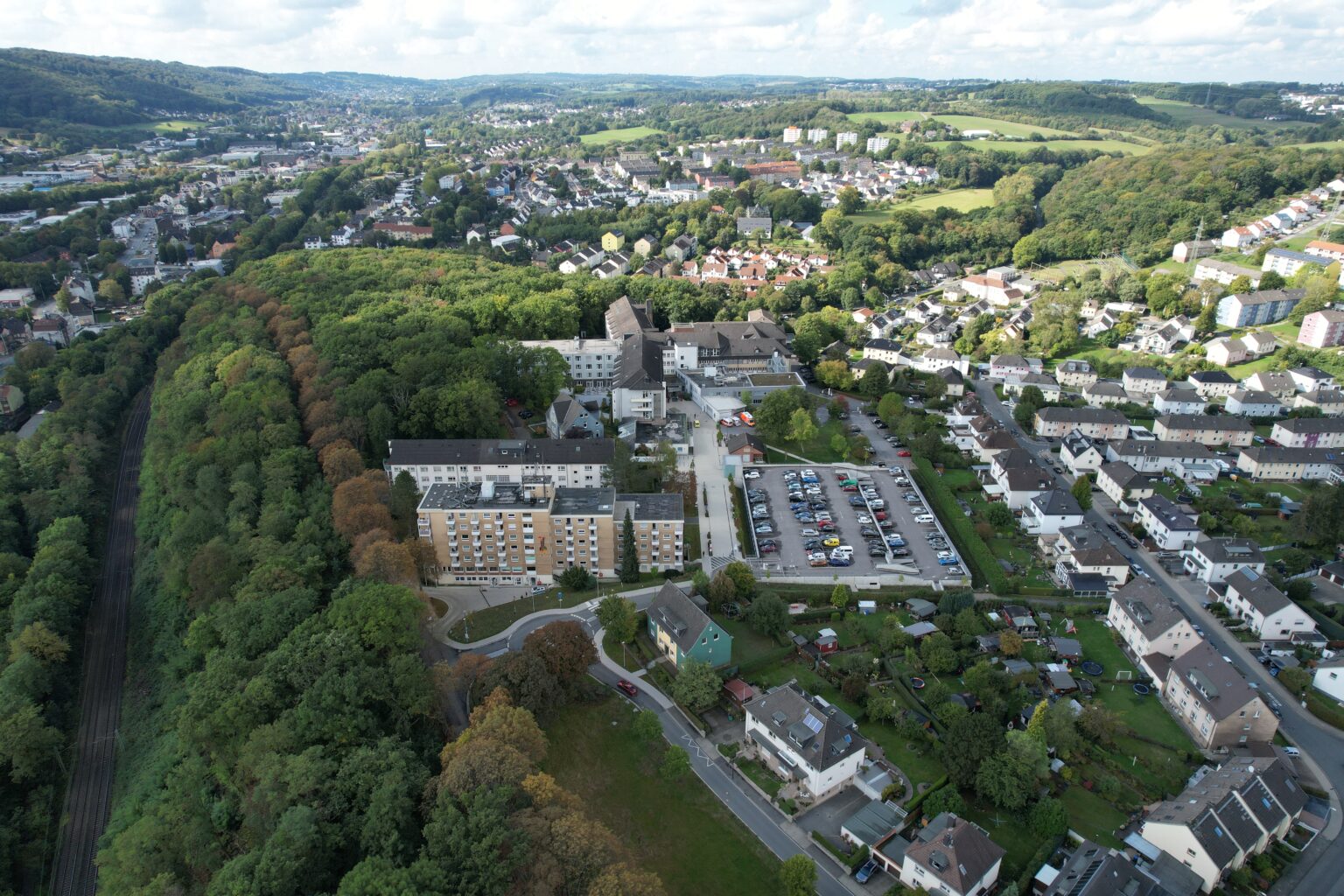 Luftaufnahme einer Stadtlandschaft mit Gebäuden, einem Parkplatz und einem bewaldeten Gebiet im Hintergrund.