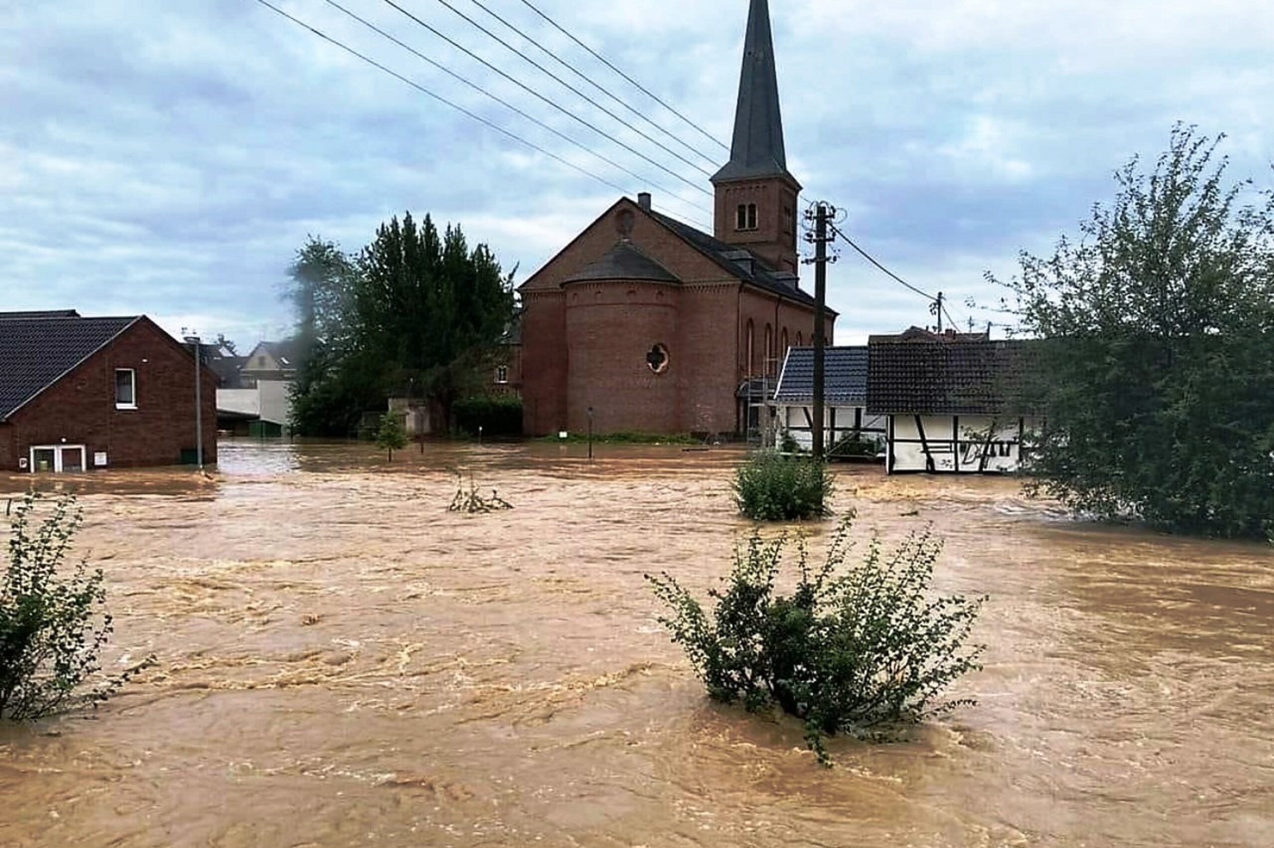 GW_Header_neu2-5120x0-c-default Überflutetes Dorf mit Kirche und Häusern bei Hochwasserereignis, Naturkatastrophe, braunes Wasser bedeckt die Straßen.