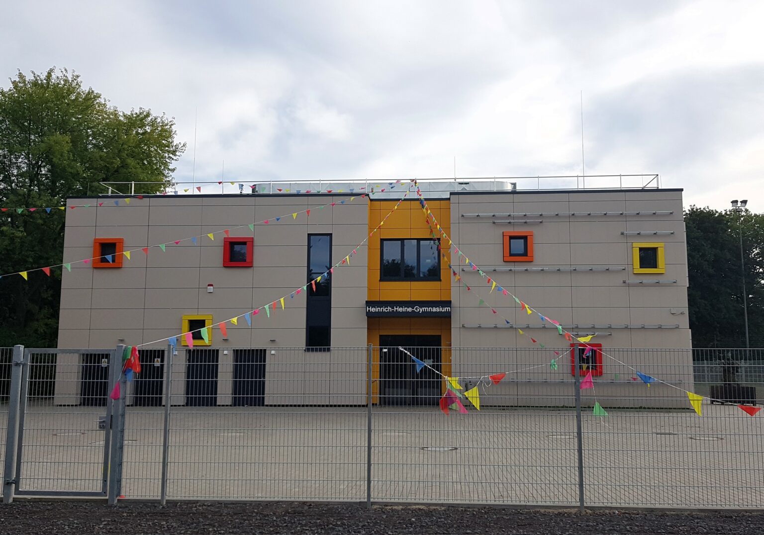HHG_4-1536x1075.2-c-default Modernes Schulgebäude mit bunten Fensterrahmen und Wimpelketten im Vordergrund an einem bewölkten Tag.