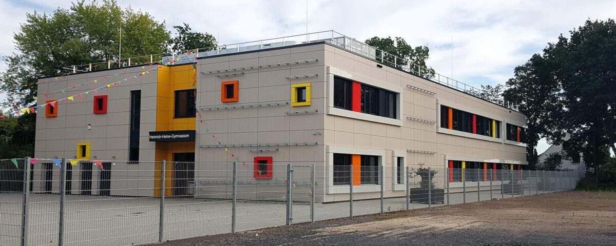 Modernes zweistöckiges Schulgebäude mit bunten Fenstern und Dekorationen, umgeben von einem Zaun und Bäumen.