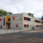 Modernes zweistöckiges Schulgebäude mit bunten Fenstern und Dekorationen, umgeben von einem Zaun und Bäumen.