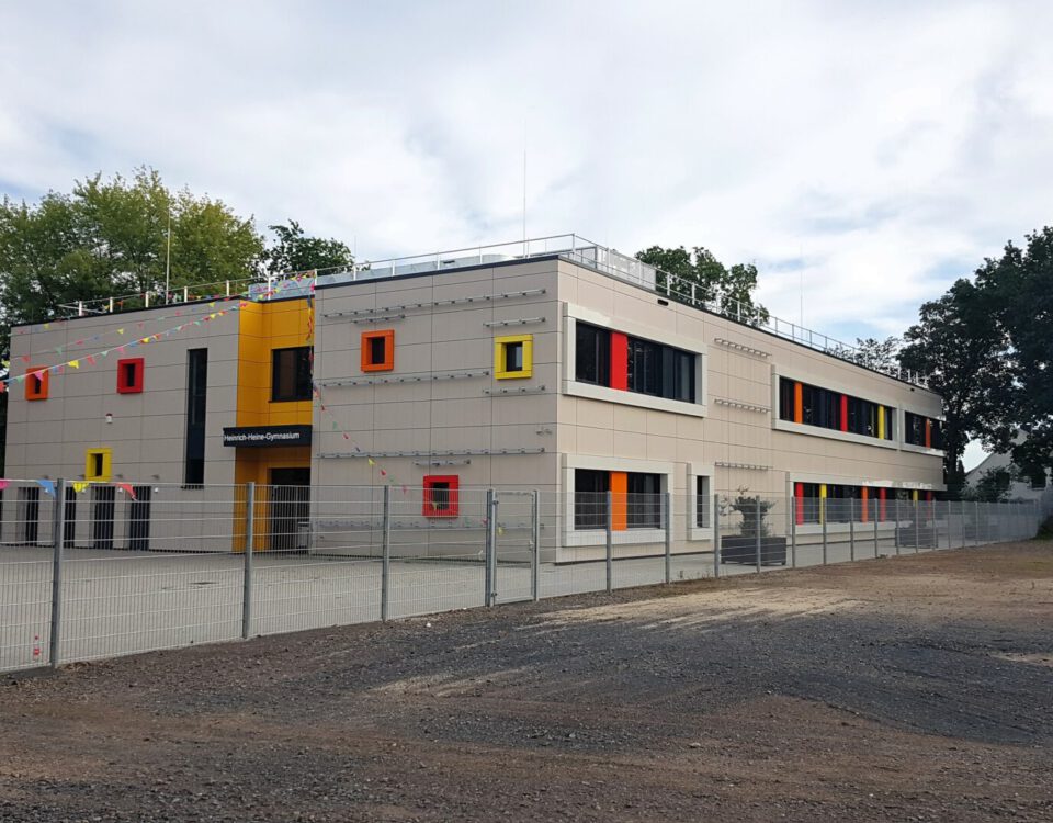 Modernes zweistöckiges Schulgebäude mit bunten Fenstern und Dekorationen, umgeben von einem Zaun und Bäumen.