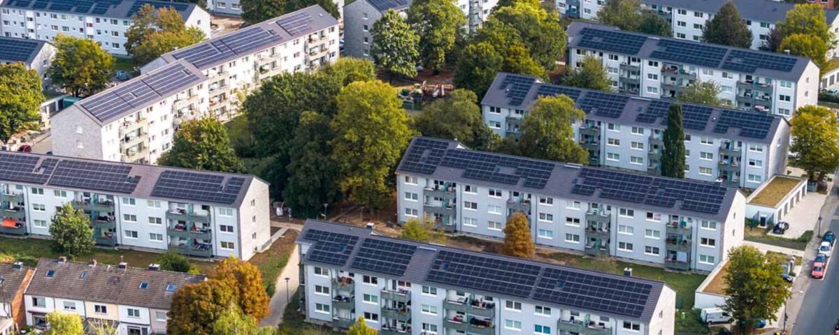 Luftaufnahme einer Wohnsiedlung mit Solaranlagen auf den Dächern, umgeben von Bäumen und parkenden Autos.