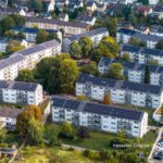 Luftaufnahme einer Wohnsiedlung mit Solaranlagen auf den Dächern, umgeben von Bäumen und parkenden Autos.