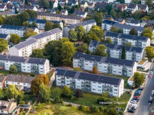 Luftaufnahme einer Wohnsiedlung mit Solaranlagen auf den Dächern, umgeben von Bäumen und parkenden Autos.