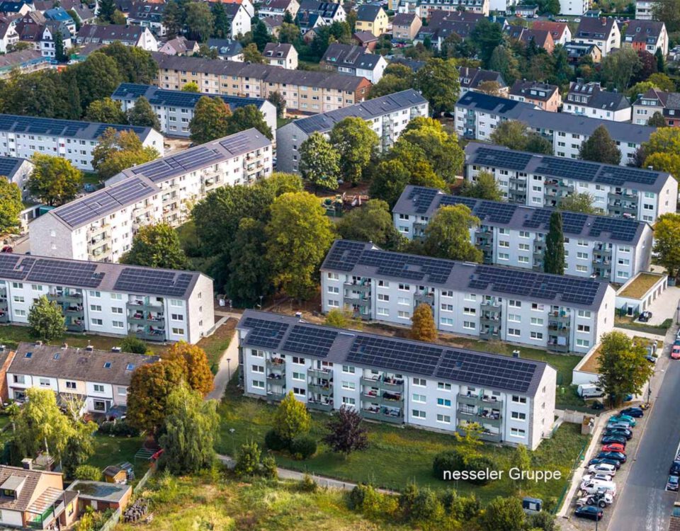 Luftaufnahme einer Wohnsiedlung mit Solaranlagen auf den Dächern, umgeben von Bäumen und parkenden Autos.