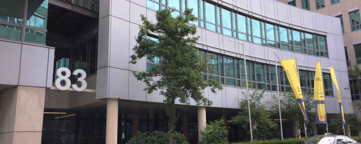 Moderne Bürogebäude mit Glasfassade, Nummer 83, gelbe Fahnen, und einem geparkten Auto im Vordergrund.