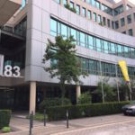 Moderne Bürogebäude mit Glasfassade, Nummer 83, gelbe Fahnen, und einem geparkten Auto im Vordergrund.