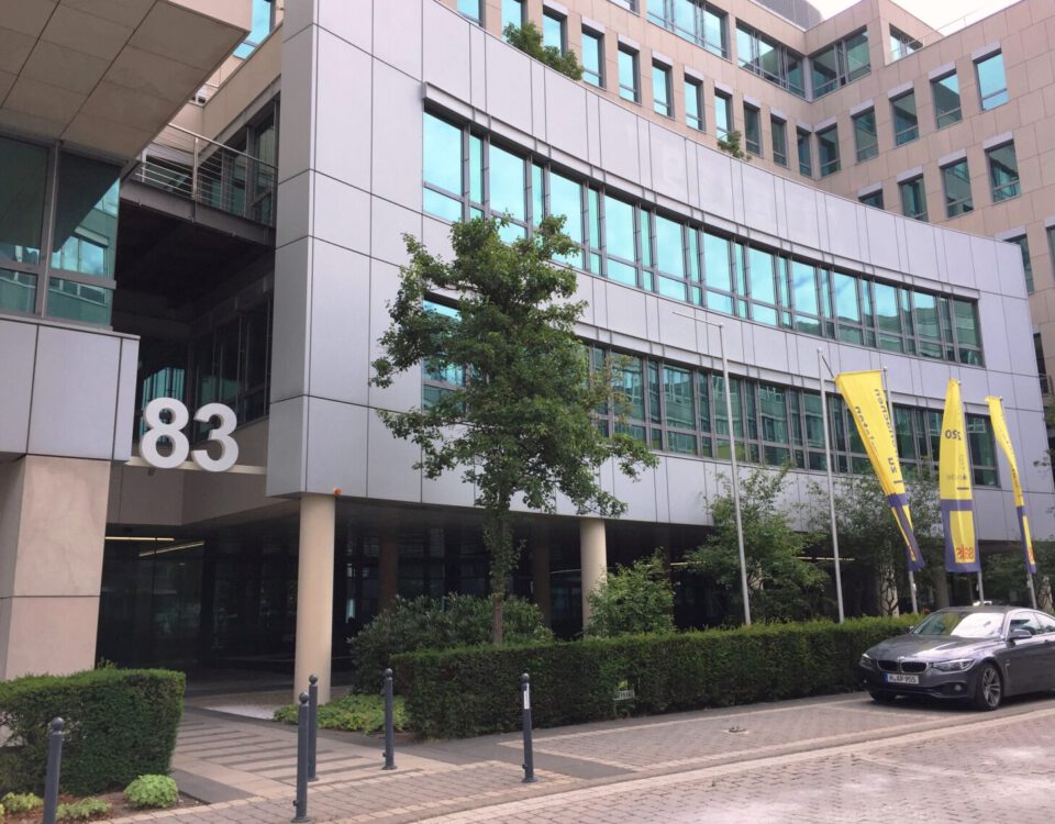 Moderne Bürogebäude mit Glasfassade, Nummer 83, gelbe Fahnen, und einem geparkten Auto im Vordergrund.