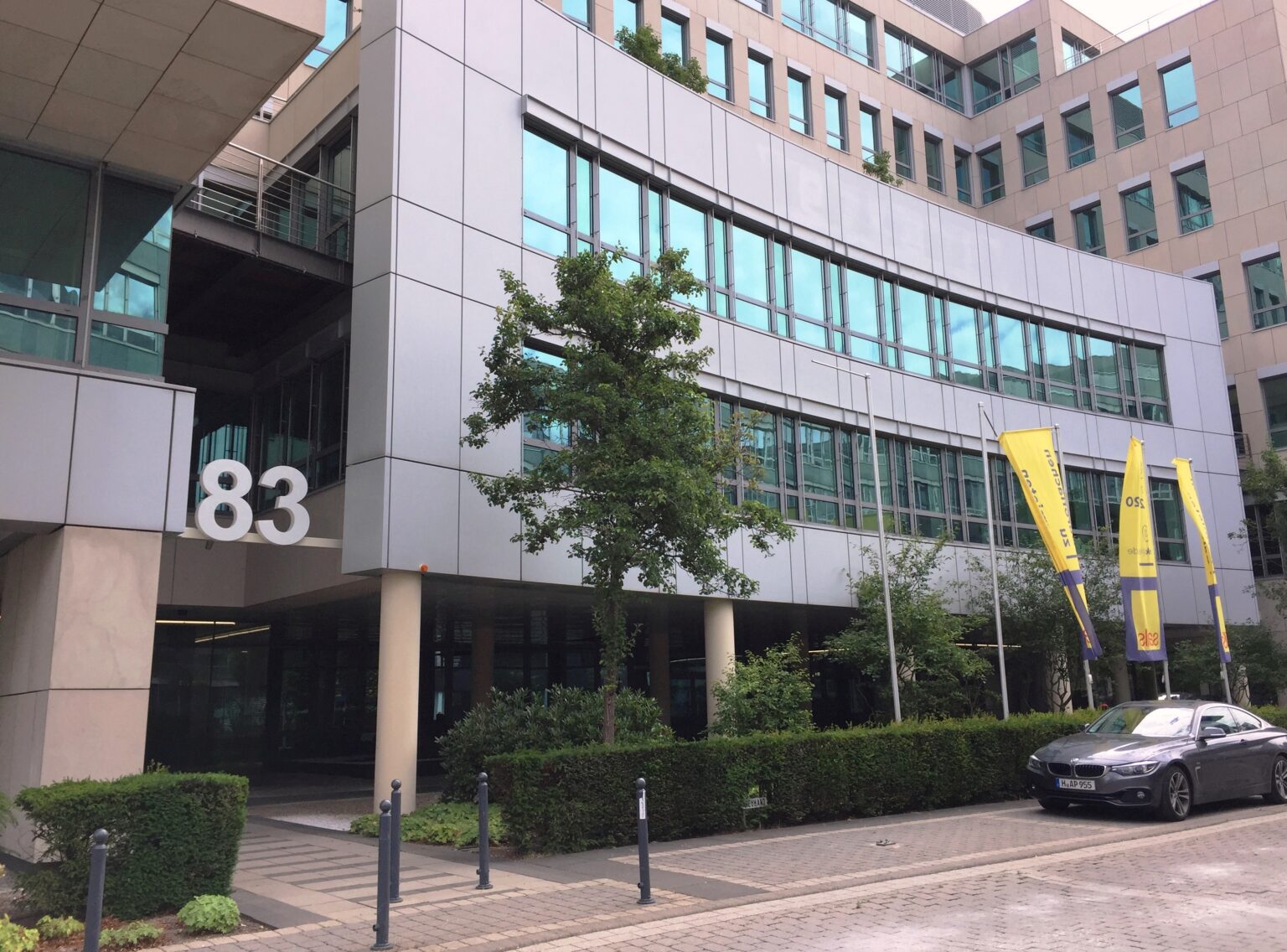 Moderne Bürogebäude mit Glasfassade, Nummer 83, gelbe Fahnen, und einem geparkten Auto im Vordergrund.