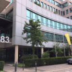 Bürogebäude mit der Nummer 83, moderne Architektur, gelbe Fahnen und parkendes Auto im Vordergrund.