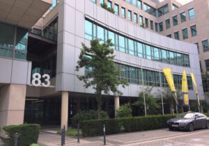 Bürogebäude mit der Nummer 83, moderne Architektur, gelbe Fahnen und parkendes Auto im Vordergrund.