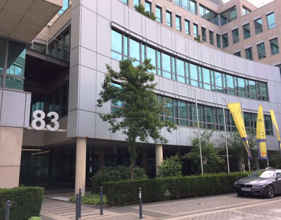 Bürogebäude mit der Nummer 83, moderne Architektur, gelbe Fahnen und parkendes Auto im Vordergrund.