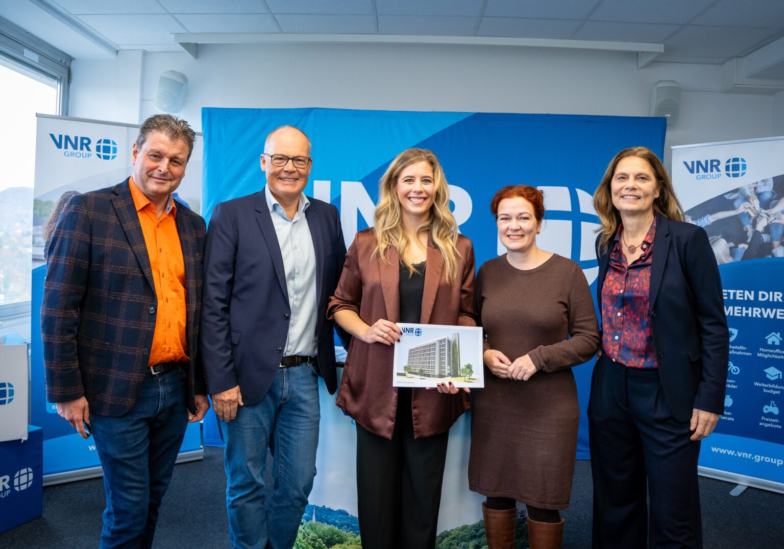 Fünf Geschäftsleute posieren vor einem Banner der VNR Group im Bürosettings für ein Firmenereignis.